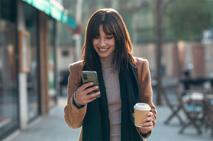 Fröhliche Frau geht mit einem Kaffe in der Hand die Straße entlang und schaut dabei auf ihr Smartphone