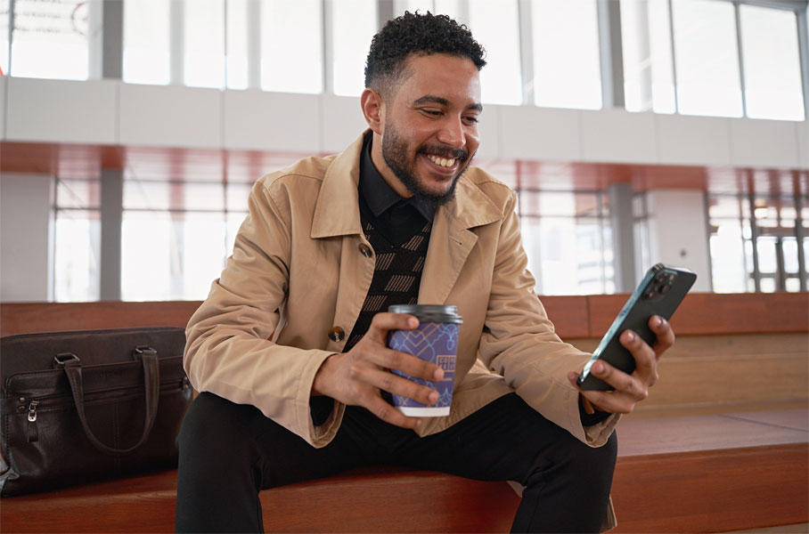Sitzender Mann blickt lachend auf sein Smartphone einen Kaffeebecher in der anderen Hand